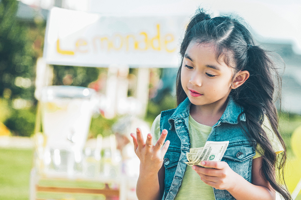 young girl counting money in front of lemonade stand