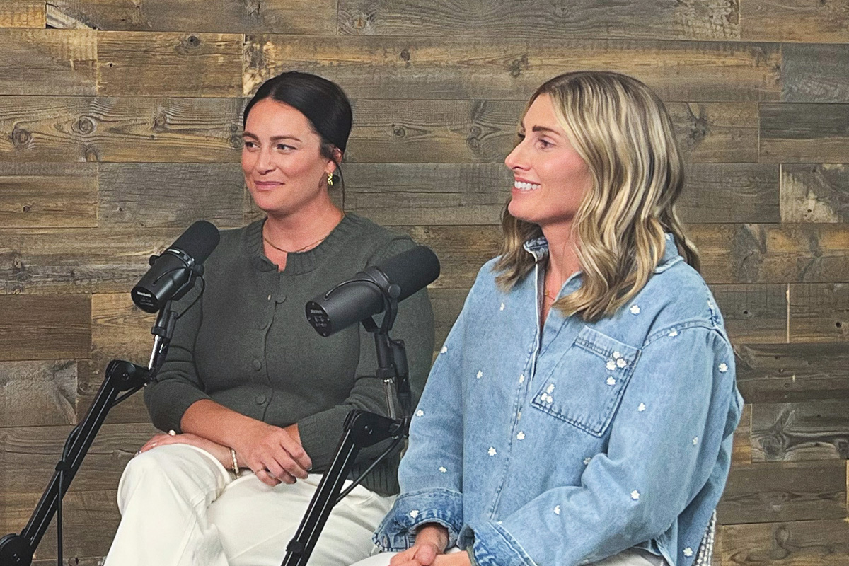 brunette woman and blonde woman sitting on stools in front of podcast mics