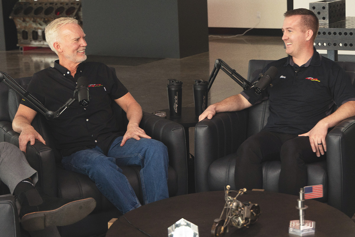 older, white-haired man and younger man in black short-sleeved shirts at the podcast mic