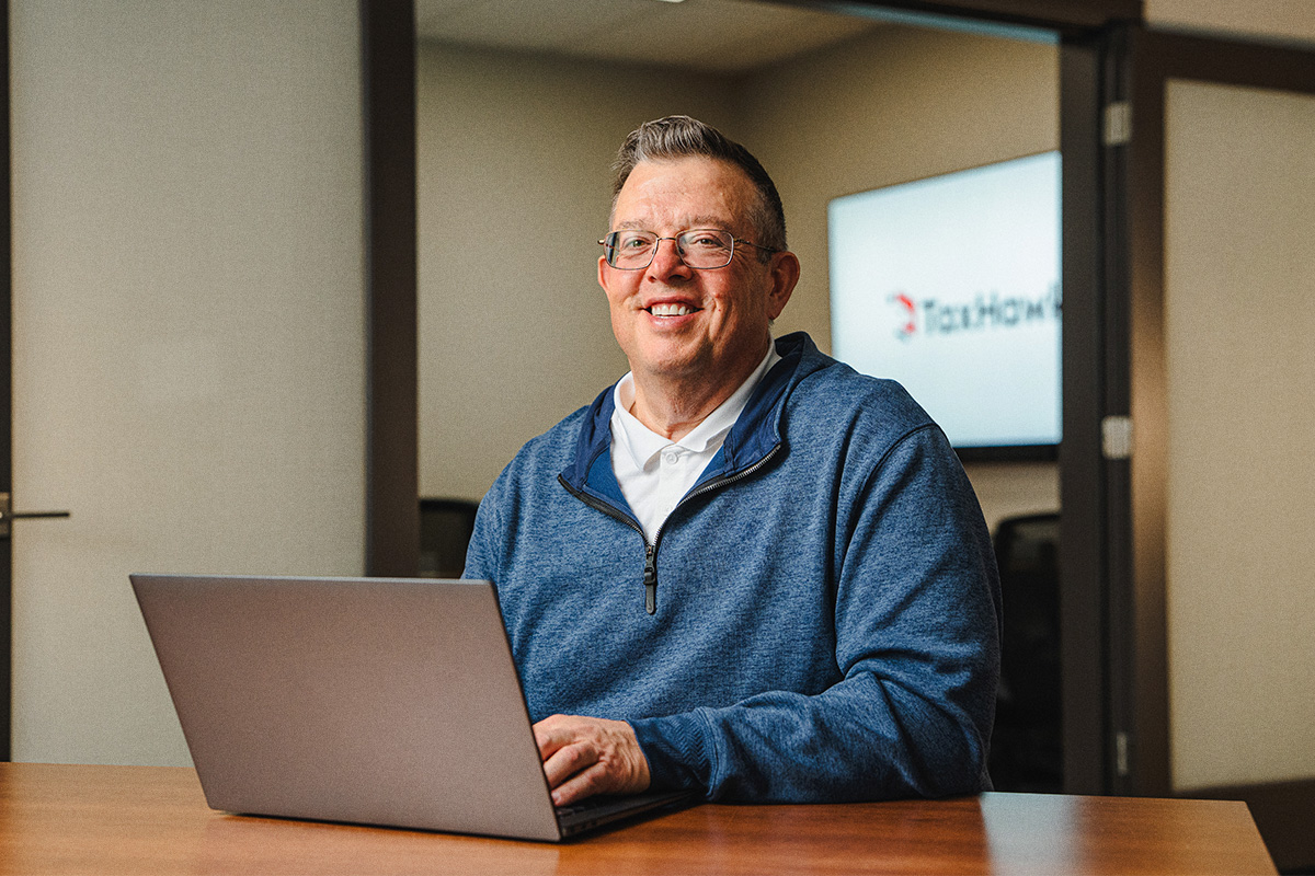 man with glasses and blue sweater smiling and sitting in front of computer