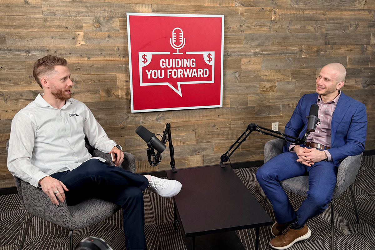 Two men sitting across a low table discussing in front of microphones