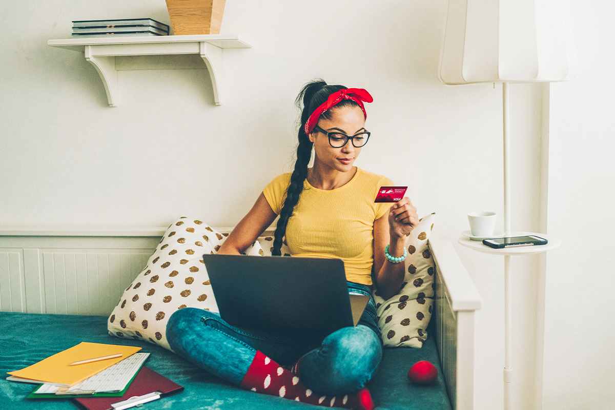 young woman sitting on her bed with her laptop while looking at her Mountain America card